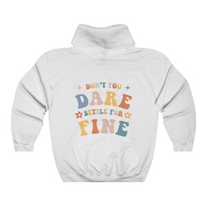 Puede incluir: Sudadera con capucha blanca con el texto "Don't you dare settle for fine" en letras retro de colores.