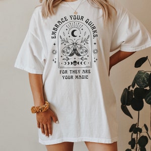 May include: White oversized t-shirt with a black graphic design featuring a moth, crescent moon, sun, and stars. The text reads "Embrace Your Quirks. For They Are Your Magic."