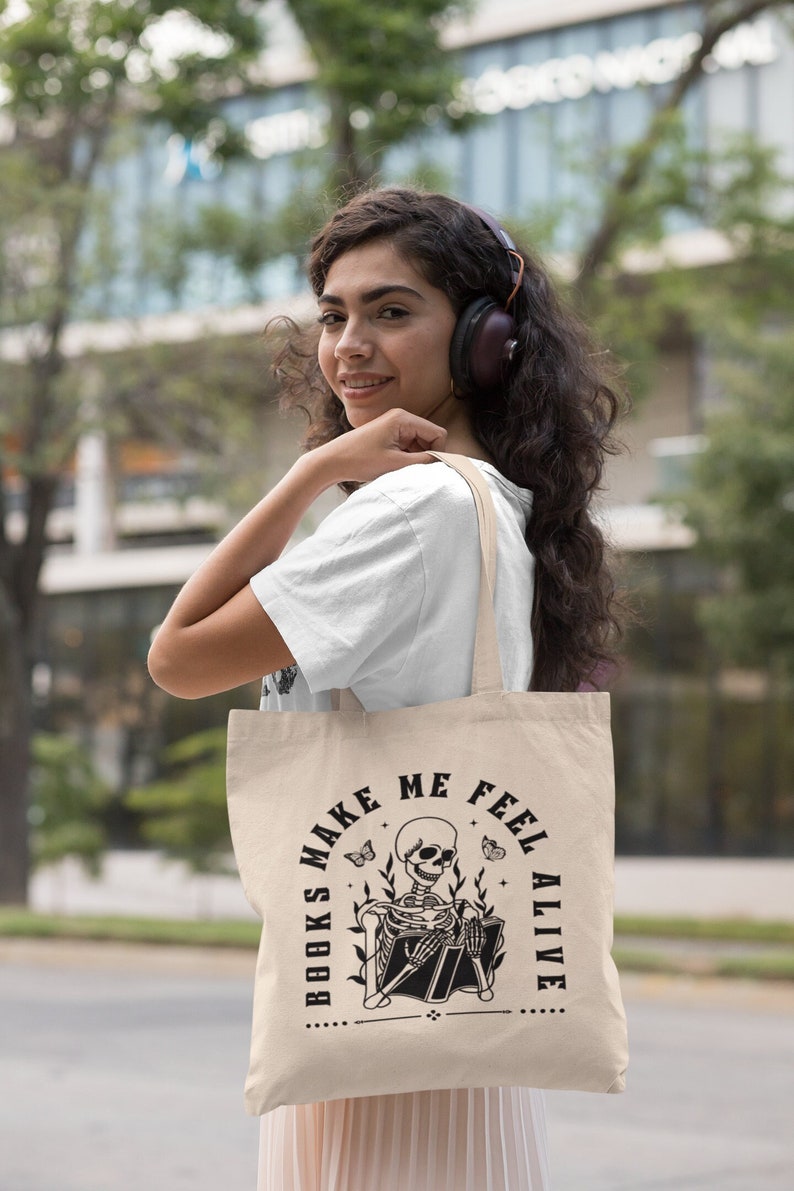Pu&ograve; includere: Una borsa di tela beige con una grafica in bianco e nero di uno scheletro che legge un libro. Il testo sulla borsa recita "Books make me feel alive".