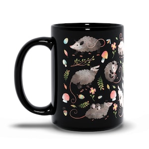May include: Black ceramic coffee mug with a white and gray opossum pattern. The opossums are surrounded by flowers, leaves, and mushrooms.
