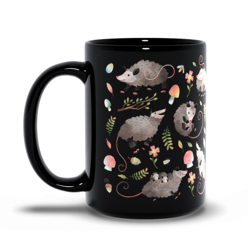 May include: Black ceramic coffee mug with a white and gray opossum pattern. The opossums are surrounded by flowers, leaves, and mushrooms.
