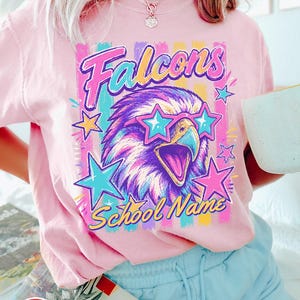May include: A light pink t-shirt featuring a graphic of a falcon wearing star-shaped sunglasses. The design includes the text "Falcons" and "School Name" in a colorful, retro-inspired font, surrounded by bright stars and vertical stripes.