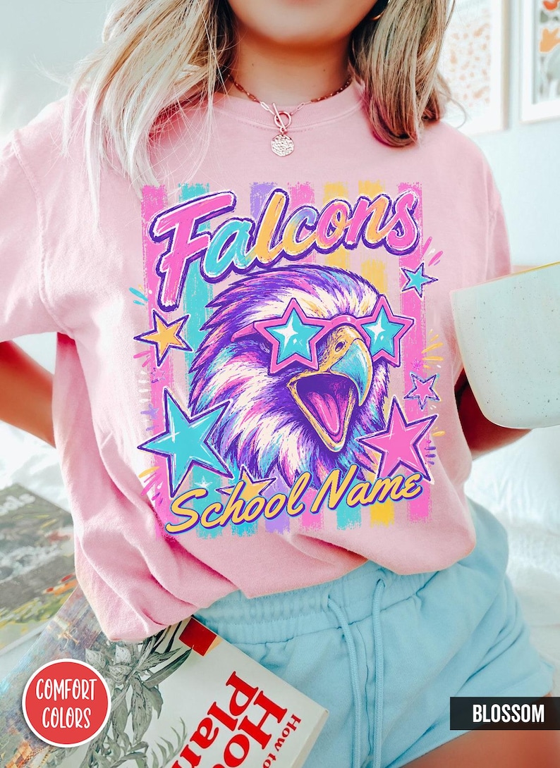 May include: A light pink t-shirt featuring a graphic of a falcon wearing star-shaped sunglasses. The design includes the text "Falcons" and "School Name" in a colorful, retro-inspired font, surrounded by bright stars and vertical stripes.
