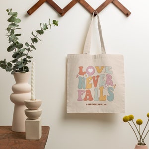 May include: A beige canvas tote bag with the words "LOVE NEVER FAILS" in rainbow colors and a heart. The text "1 Corinthians 13:8" is printed below the words.