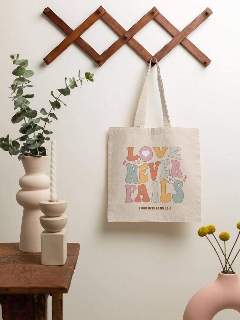 May include: A beige canvas tote bag with the words "LOVE NEVER FAILS" in rainbow colors and a heart. The text "1 Corinthians 13:8" is printed below the words.