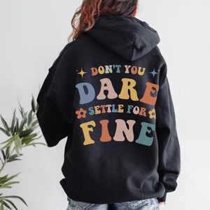 Puede incluir: Sudadera con capucha negra con el texto "Don't you dare settle for fine" en letras de colores en la espalda.