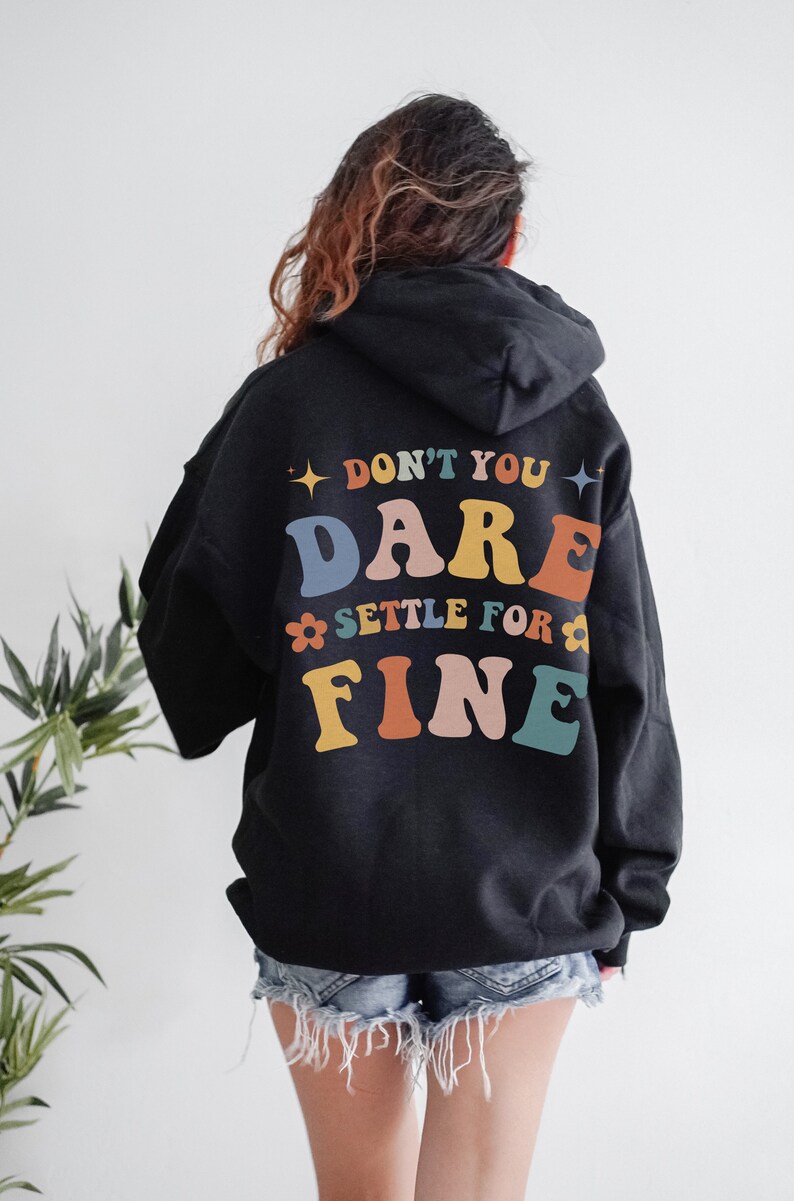 Puede incluir: Sudadera con capucha negra con el texto "Don't you dare settle for fine" en letras de colores en la espalda.