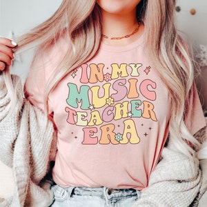 以下が含まれることがあります： レトロなフォントでカラフルなアクセントと花がついた「In My Music Teacher Era」というテキストが書かれたピンクのTシャツです。