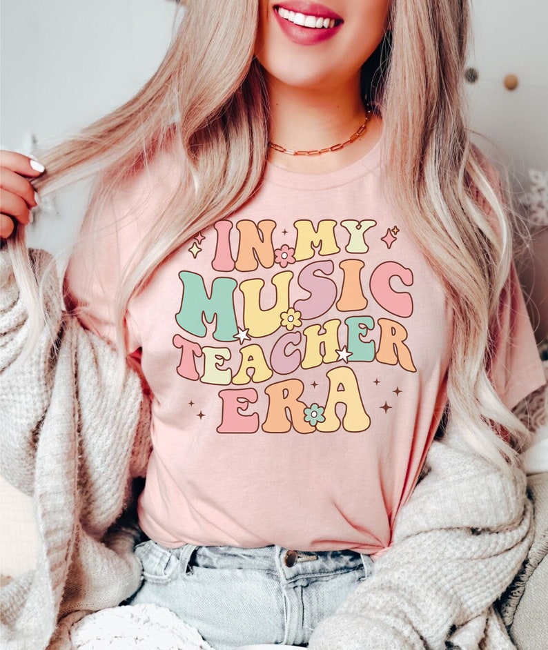 以下が含まれることがあります： レトロなフォントでカラフルなアクセントと花がついた「In My Music Teacher Era」というテキストが書かれたピンクのTシャツです。