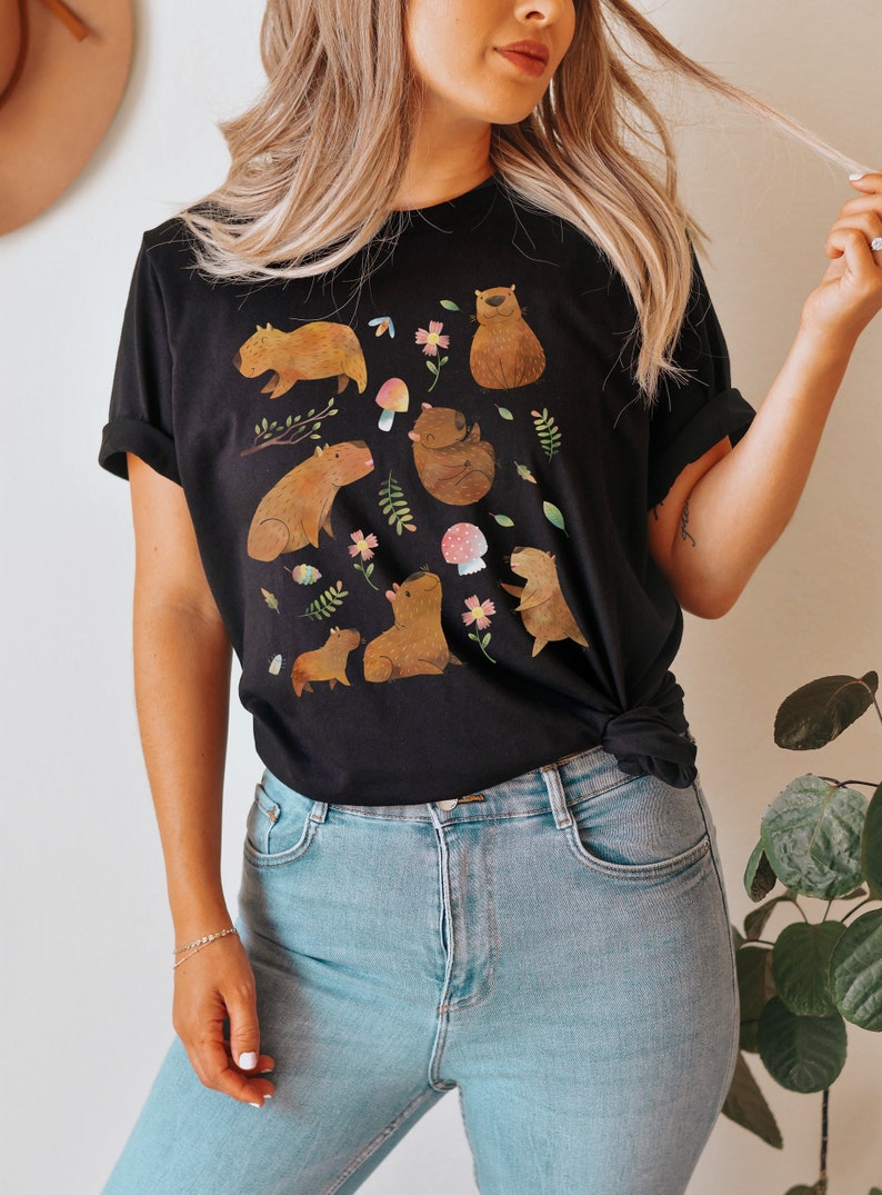 May include: A black t-shirt with a graphic print of multiple capybaras in a forest setting. The capybaras are brown and have white accents. The forest setting includes green leaves, flowers, and mushrooms.
