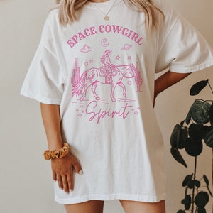 May include: White oversized t-shirt with a pink graphic of a cowgirl riding a horse in space. The graphic includes stars, planets, clouds, and cacti. The text "Space Cowgirl Spirit" is printed on the shirt.