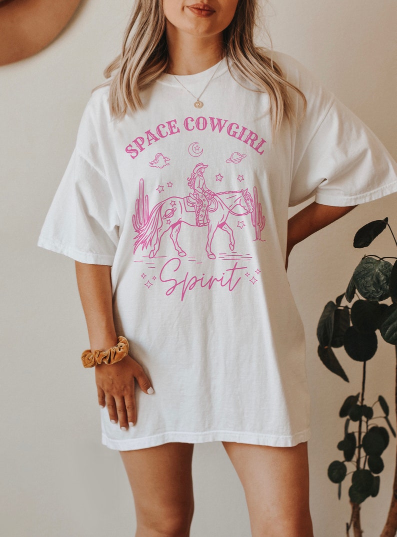 May include: White oversized t-shirt with a pink graphic of a cowgirl riding a horse in space. The graphic includes stars, planets, clouds, and cacti. The text "Space Cowgirl Spirit" is printed on the shirt.