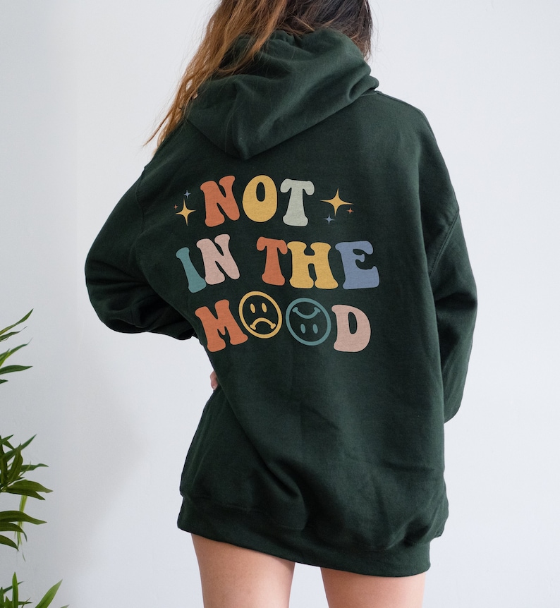 Pu&ograve; includere: Una felpa con cappuccio verde scuro con il testo "NOT IN THE MOOD" in colori arcobaleno con emoji di facce tristi.