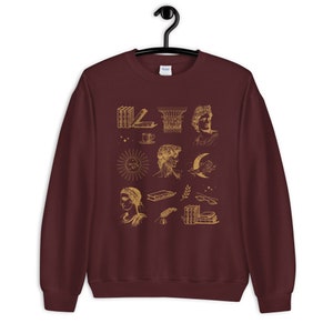 Puede incluir: Una sudadera color burdeos con un estampado dorado de varias ilustraciones, incluyendo libros, un sol, una luna, una columna y una taza de caf&eacute;.