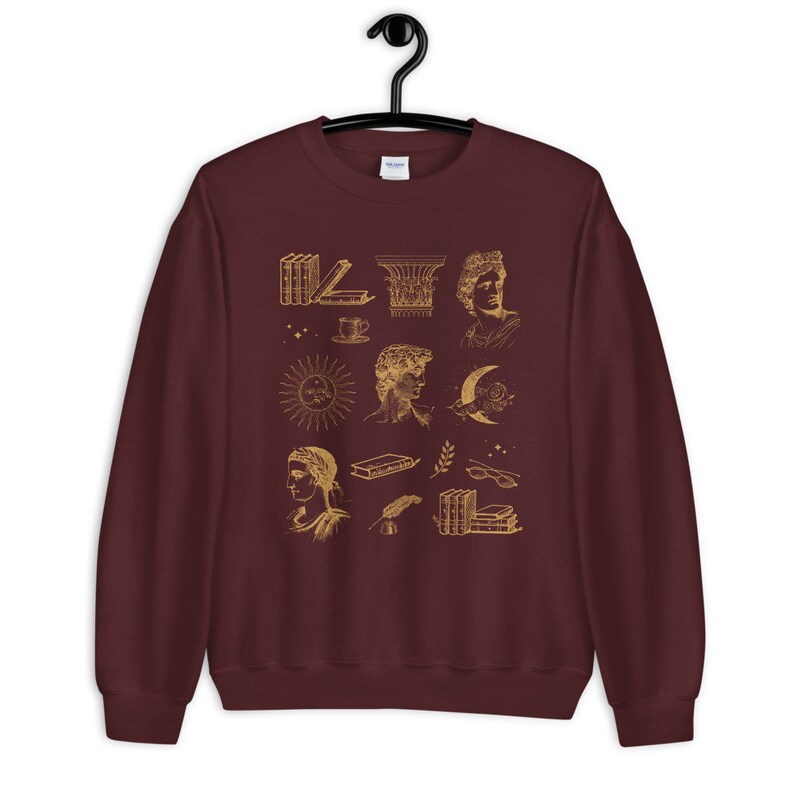 Puede incluir: Una sudadera color burdeos con un estampado dorado de varias ilustraciones, incluyendo libros, un sol, una luna, una columna y una taza de caf&eacute;.