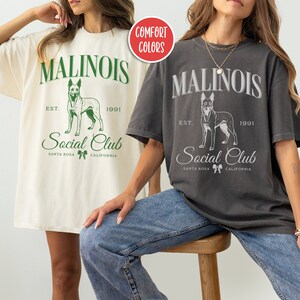 May include: Two women wearing oversized t-shirts with a graphic of a dog and the text "Malinois Social Club Est. 1991 Santa Rosa California". The t-shirt on the left is cream colored with green text and the t-shirt on the right is charcoal gray with white text.