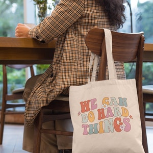 Op de afbeelding: Een witte canvas tote bag met de tekst "WE CAN DO HARD THINGS" in kleurrijke retro-stijl letters met kleine bloem accenten.