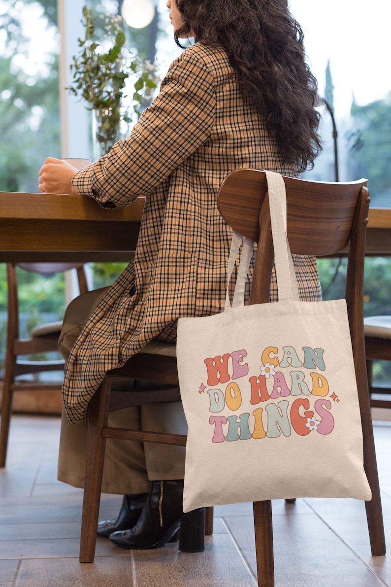 Op de afbeelding: Een witte canvas tote bag met de tekst "WE CAN DO HARD THINGS" in kleurrijke retro-stijl letters met kleine bloem accenten.