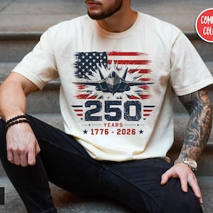 May include: Ivory-colored t-shirt featuring a graphic of an American flag, a jet, and the text "250 YEARS 1776-2026." The design incorporates red, white, and blue colors. The shirt is a Comfort Colors brand.