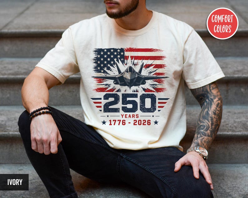 May include: Ivory-colored t-shirt featuring a graphic of an American flag, a jet, and the text "250 YEARS 1776-2026." The design incorporates red, white, and blue colors. The shirt is a Comfort Colors brand.