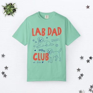 Puede incluir: Una camiseta verde claro con el texto rojo LAB DAD CLUB e ilustraciones azules de dos perros Labrador, una pelota, un juguete y huellas. La camiseta tambi&eacute;n incluye el texto azul home is where my labrador is y la inscripci&oacute;n est. 2026.