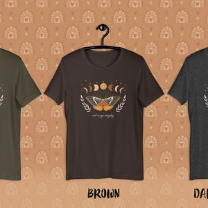 K&ouml;nnte beinhalten: Drei T-Shirts in verschiedenen Farben: Armeegr&uuml;n, Braun und Dunkelgrau meliert. Jedes Shirt zeigt ein Grafikdesign mit einem Schmetterling mit Mondphasen und dem Text "and magic unfolding".