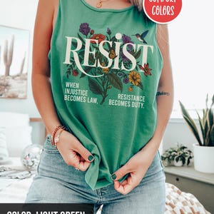 May include: A light green tank top with the word "RESIST" printed in white with a floral design and the text "When injustice becomes law, resistance becomes duty."