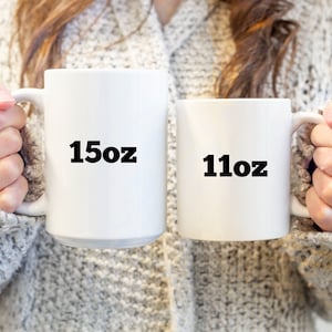 May include: Two white ceramic mugs with black text. The larger mug says "15oz" and the smaller mug says "11oz".