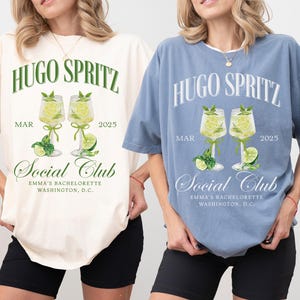May include: Two t-shirts with a graphic design of two glasses of Hugo Spritz, with the text "Hugo Spritz" and "Social Club" on the front. The t-shirts are in ivory and blue jean colors.