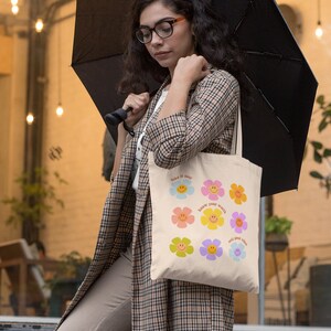 May include: A woman is holding a beige tote bag with a colorful floral design and the text "take it easy", "know your worth", and "only good vibes".