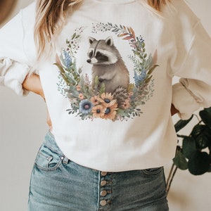 May include: White sweatshirt featuring a watercolor illustration of a raccoon surrounded by a floral wreath. The raccoon is gray and white, and the wreath includes blue, orange, and green flowers and foliage. The sweatshirt is paired with blue jeans.