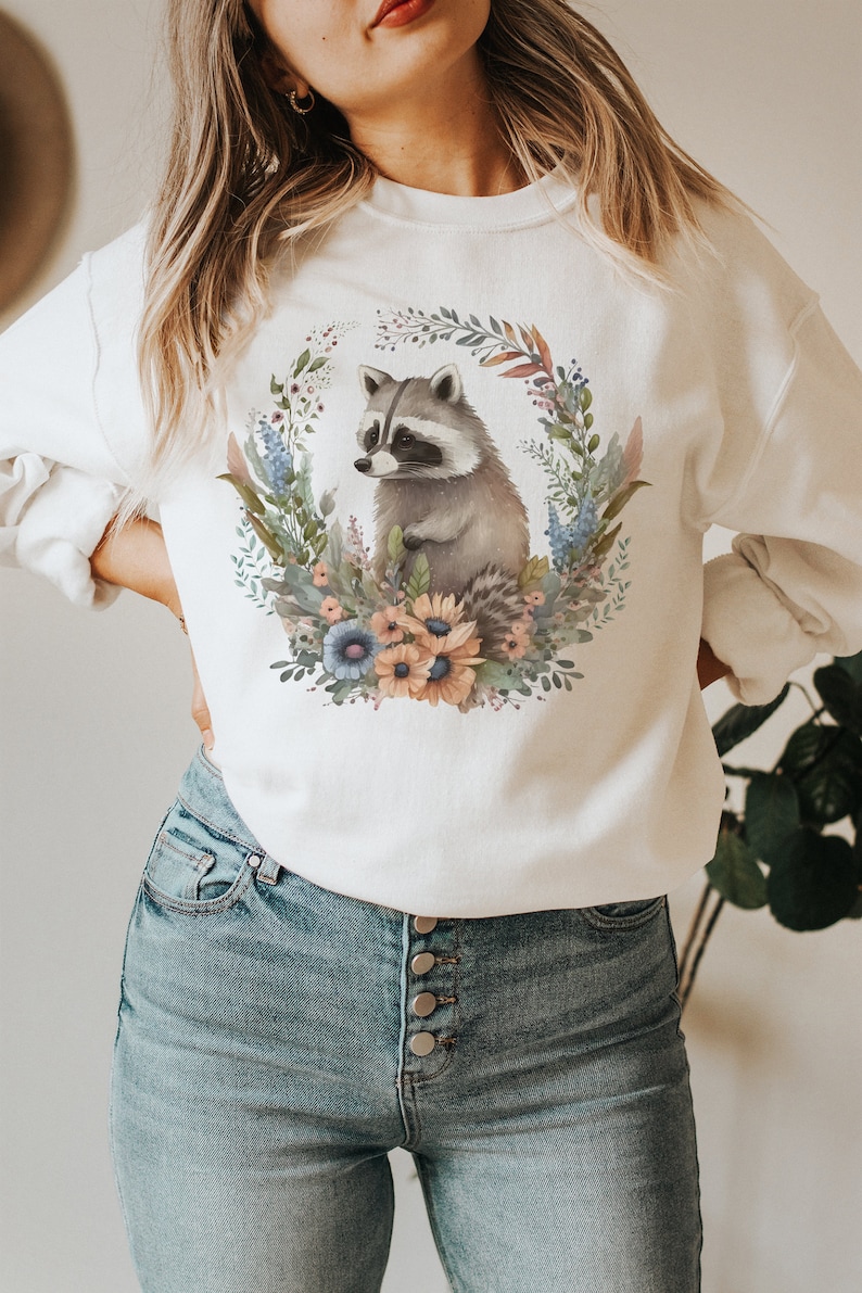 May include: White sweatshirt featuring a watercolor illustration of a raccoon surrounded by a floral wreath. The raccoon is gray and white, and the wreath includes blue, orange, and green flowers and foliage. The sweatshirt is paired with blue jeans.