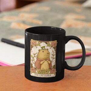 May include: Black ceramic mug with a white illustration of a capybara wearing a top hat and a red cape. The illustration is surrounded by flowers and the text "THE CAPYBARA".