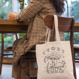 K&ouml;nnte beinhalten: Eine wei&szlig;e Tragetasche mit einer schwarz-wei&szlig;en Illustration von Pilzen und einem Halbmond-Design. Die Tasche h&auml;ngt an einem Holzstuhl.