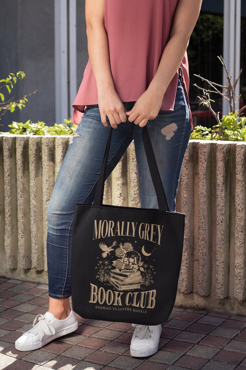 May include: A black tote bag with a gold design that says "Morally Grey Book Club" and "Enemies to Lovers Weekly". The design also features a skull, roses, and a bird.