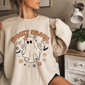 May include: A beige sweatshirt with a graphic of a ghost with the text "Spooky Season" in orange and white letters. The ghost is wearing a peace sign necklace and has flowers around its head.