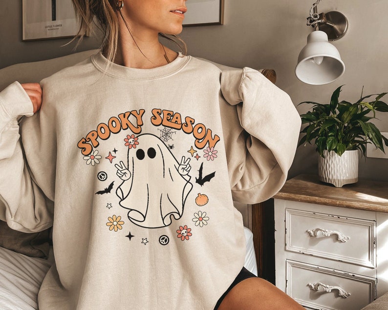 May include: A beige sweatshirt with a graphic of a ghost with the text "Spooky Season" in orange and white letters. The ghost is wearing a peace sign necklace and has flowers around its head.