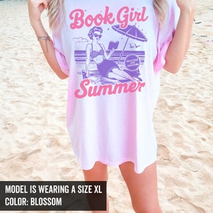 K&ouml;nnte beinhalten: Ein pink-wei&szlig;es Batik-T-Shirt mit dem Text "Book Girl Summer" in Pink und Lila. Das Design zeigt eine Comic-Frau im Badeanzug, die ein Buch am Strand mit einem Sonnenschirm liest, und den Text "Chapters and Sunshine".