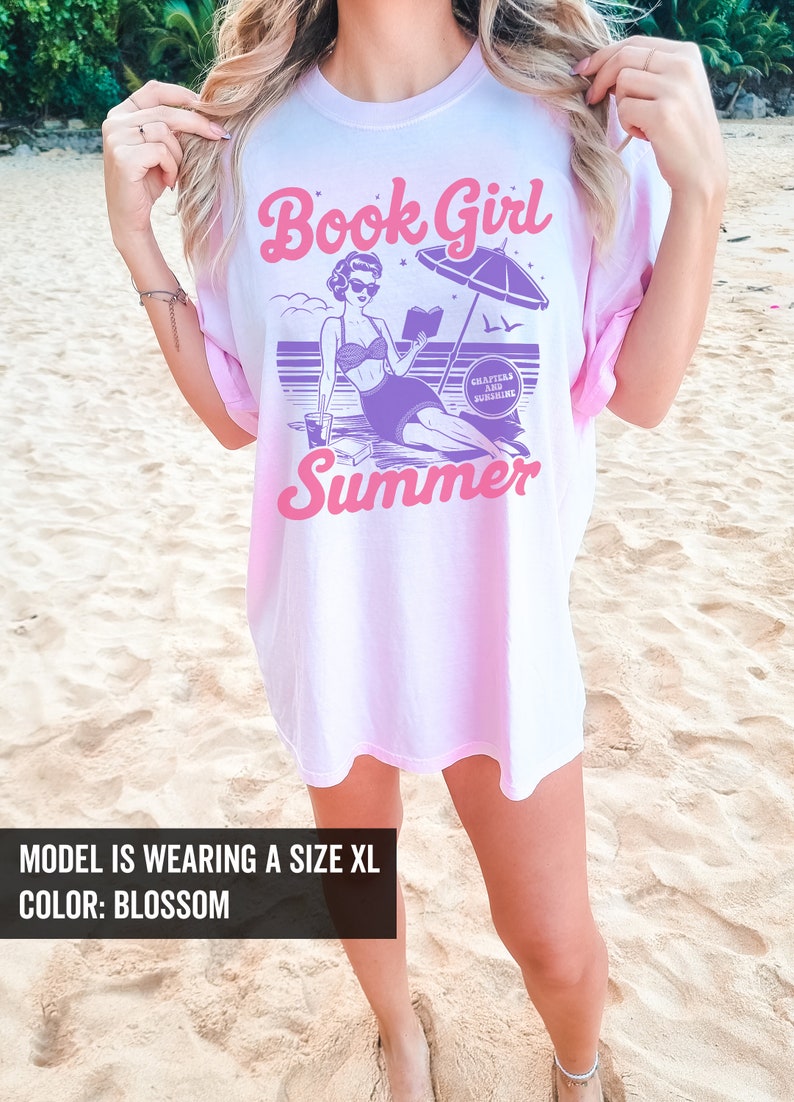 K&ouml;nnte beinhalten: Ein pink-wei&szlig;es Batik-T-Shirt mit dem Text "Book Girl Summer" in Pink und Lila. Das Design zeigt eine Comic-Frau im Badeanzug, die ein Buch am Strand mit einem Sonnenschirm liest, und den Text "Chapters and Sunshine".