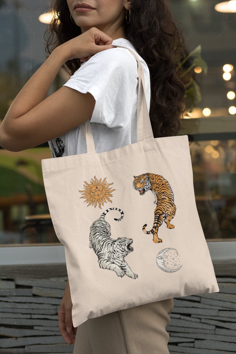 Op de afbeelding: Een beige tote bag met een witte tijger, een oranje tijger, een zon en een maanontwerp. De tas heeft twee hengsels.