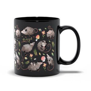 May include: Black coffee mug with a white and gray opossum pattern. The opossums are surrounded by flowers and mushrooms.