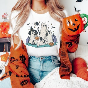 May include: White t-shirt with a graphic of five cartoon cats dressed in Halloween costumes. The cats are holding pumpkins and candy bags. The graphic is set against a dark blue background with stars and a crescent moon.