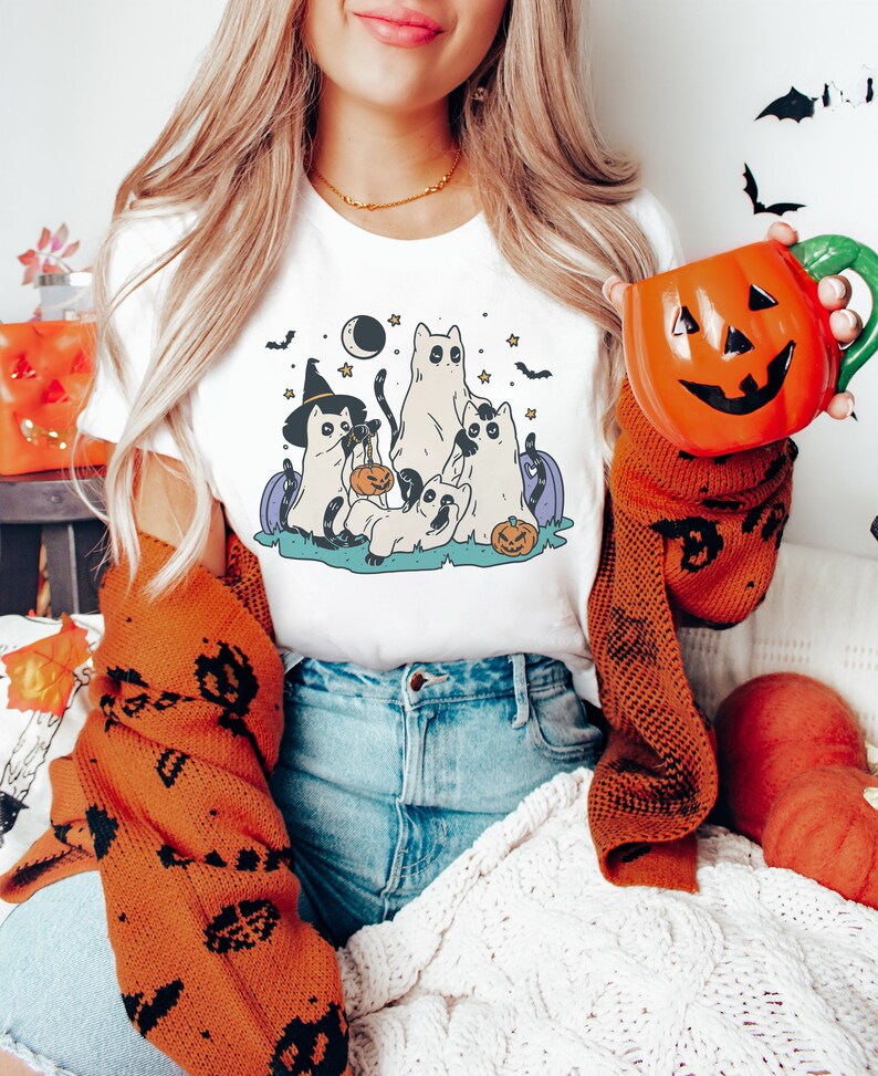 May include: White t-shirt with a graphic of five cartoon cats dressed in Halloween costumes. The cats are holding pumpkins and candy bags. The graphic is set against a dark blue background with stars and a crescent moon.