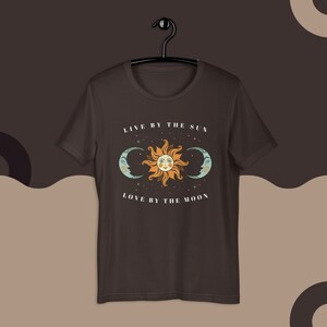 May include: A dark brown t-shirt with a white graphic of a sun and two moons. The text "Live by the sun, love by the moon" is printed on the shirt.