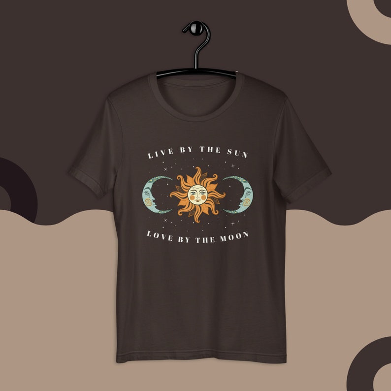 May include: A dark brown t-shirt with a white graphic of a sun and two moons. The text "Live by the sun, love by the moon" is printed on the shirt.