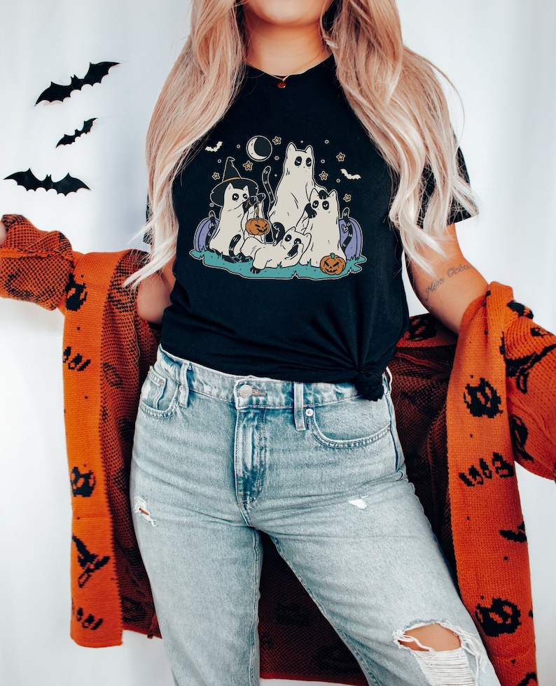 May include: Black t-shirt with a graphic of four cats dressed in Halloween costumes. The cats are wearing witch hats, ghost costumes, and holding a pumpkin. The graphic is surrounded by stars and bats.