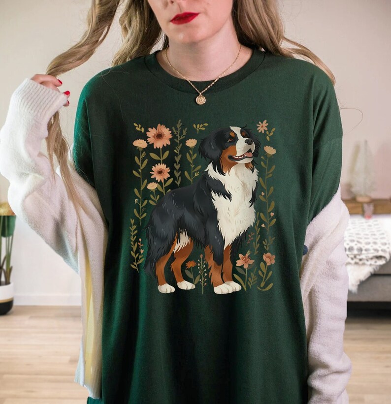 Pu&ograve; includere: Una maglietta verde con un'illustrazione di un cane da montagna bernese circondato da fiori. Il cane &egrave; marrone, nero e bianco con uno sfondo floreale.