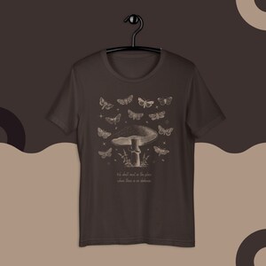 May include: A dark brown t-shirt with a vintage illustration of a mushroom and butterflies. The text "We shall meet in the place where there is no darkness" is printed below the image.