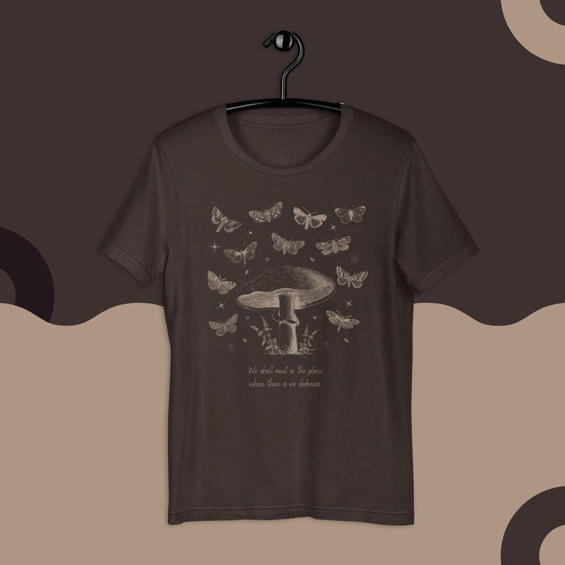 May include: A dark brown t-shirt with a vintage illustration of a mushroom and butterflies. The text "We shall meet in the place where there is no darkness" is printed below the image.