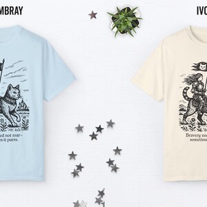 Peut inclure: Deux t-shirts avec un imprim&eacute; graphique d'une femme chevauchant un chat. Le texte sur le t-shirt est "Bravery need not roar - sometimes it purrs." Le t-shirt de gauche est bleu clair et le t-shirt de droite est ivoire.